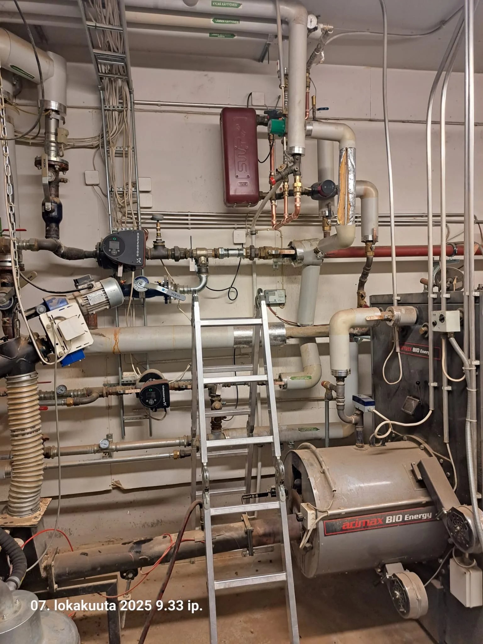 Kuva näyttää lämmityskattilahuoneen, jossa on putkia, pumppuja, venttiileitä ja kattila. Metalliset tikkaat ovat huoneen keskellä. Valo on himmeää ja teknistä.