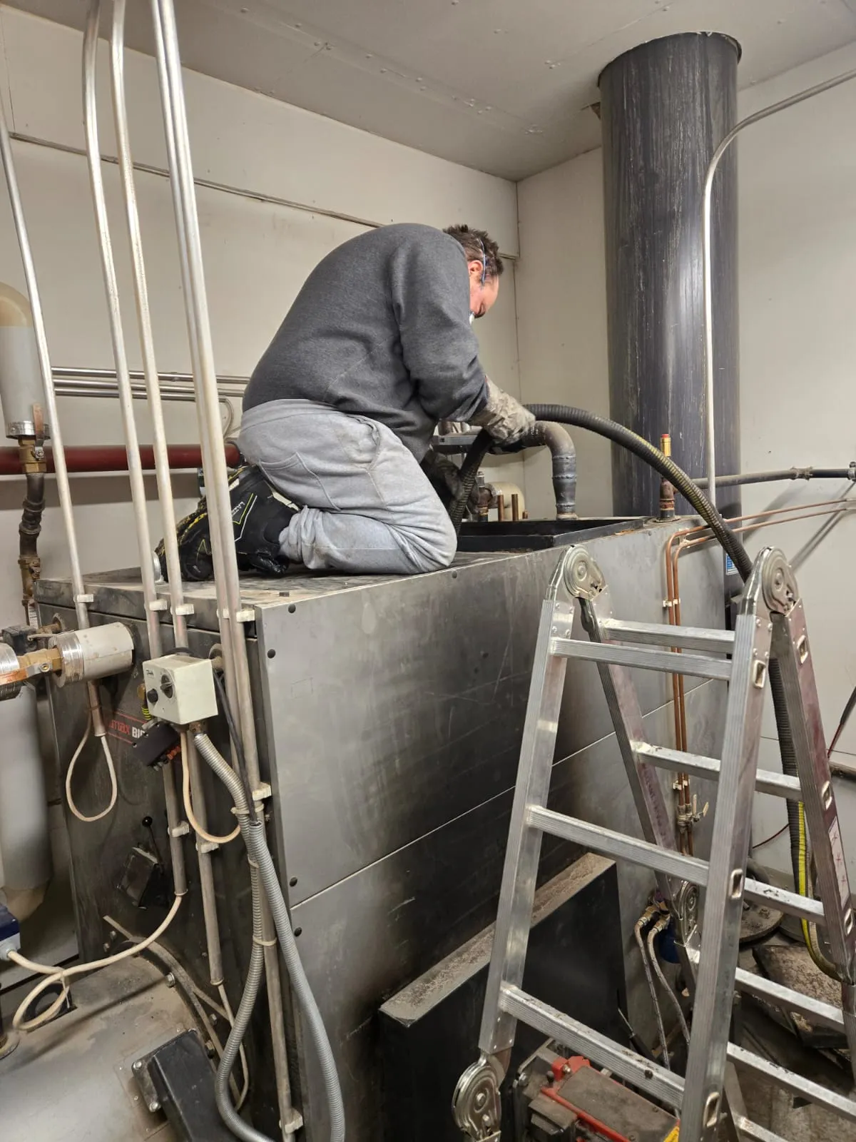 Mies polvillaan metallisella lämmityskattilalla, käyttäen letkua, vieressä tikkaat, harmaat vaatteet, sisätiloissa, kirkas valaistus.