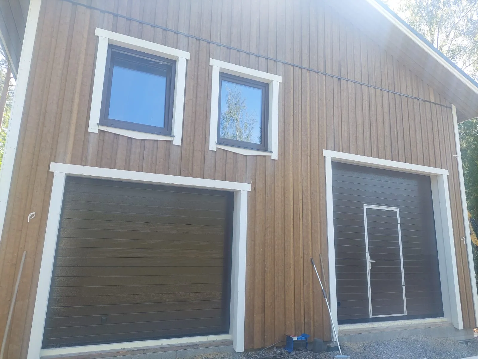 Ruskea puurakennus, jossa on kaksi autotallin ovea ja kaksi ikkunaa valkoisilla kehyksillä, aurinkoisessa säässä.