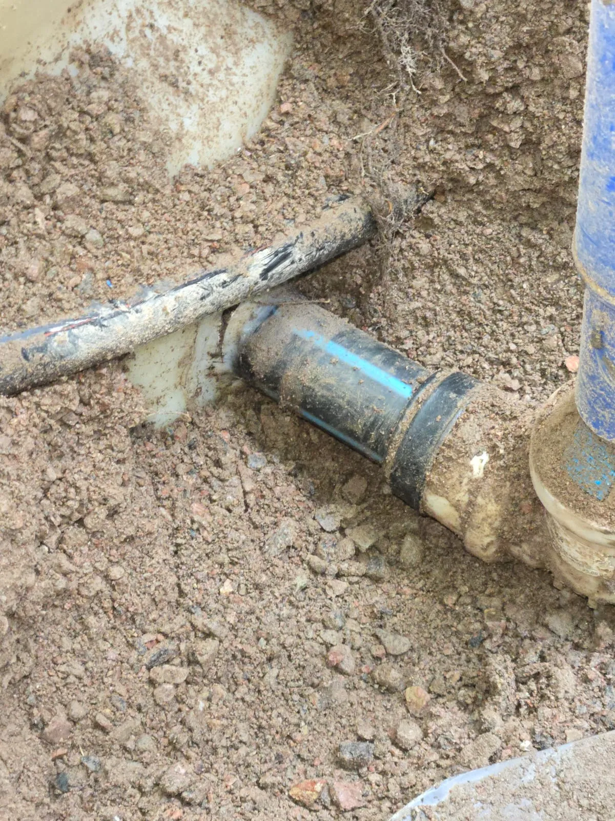 Maaperään kaivettu musta ja sininen muoviputki, jossa on liitoskohta ja valkoinen muovipala, valaistus on luonnollinen ja hieman samea.